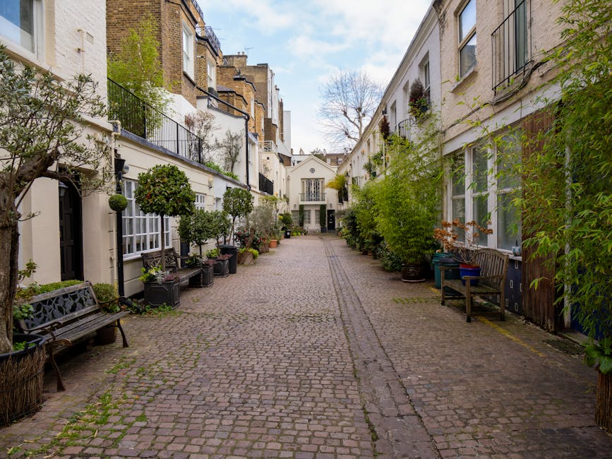 A cobblestone alleyway in an urban residential area with white-painted terraced buildings on both sides, featuring large windows with black metal railings and flower boxes. The alley is lined with pot