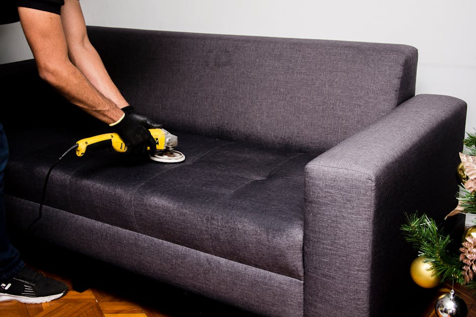 A person wearing black gloves operates a handheld electric upholstery cleaner on a dark gray fabric sofa in a well-lit living room. The sofa has a modern design with a straight backrest and armrests,
